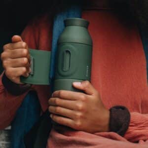 A person unscrews the lid of a green Closca Wave – Amazonia reusable water bottle, wearing a red long-sleeve shirt and blue rucksack—highlighting innovative eco-friendly finds at your favourite gift shop. The hands and bottle are the main focus. Gate21.ee