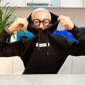 A person with short blond hair and round glasses pulls the collar of a NOFUCKSGIVEN Hoodie over their mouth and nose at a table. Green plants and blue bags featuring Estonian design appear in the background. Gate21.ee