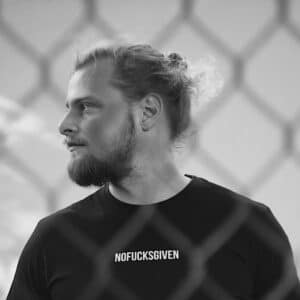 A man with a beard and long hair tied back looks to his left, wearing a black T-shirt with NOFUCKSGIVEN printed on it. A chain-link fence is visible in the foreground. The black-and-white photo captures curated Estonian design attitude. Gate21.ee