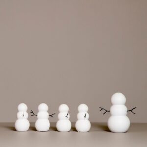 SNOWMAN features four minimalist snowman figures of different sizes in a row, each crafted from smooth white spheres with black stick arms, evoking curated d&eacute;cor vibes against a plain beige background. Gate21.ee