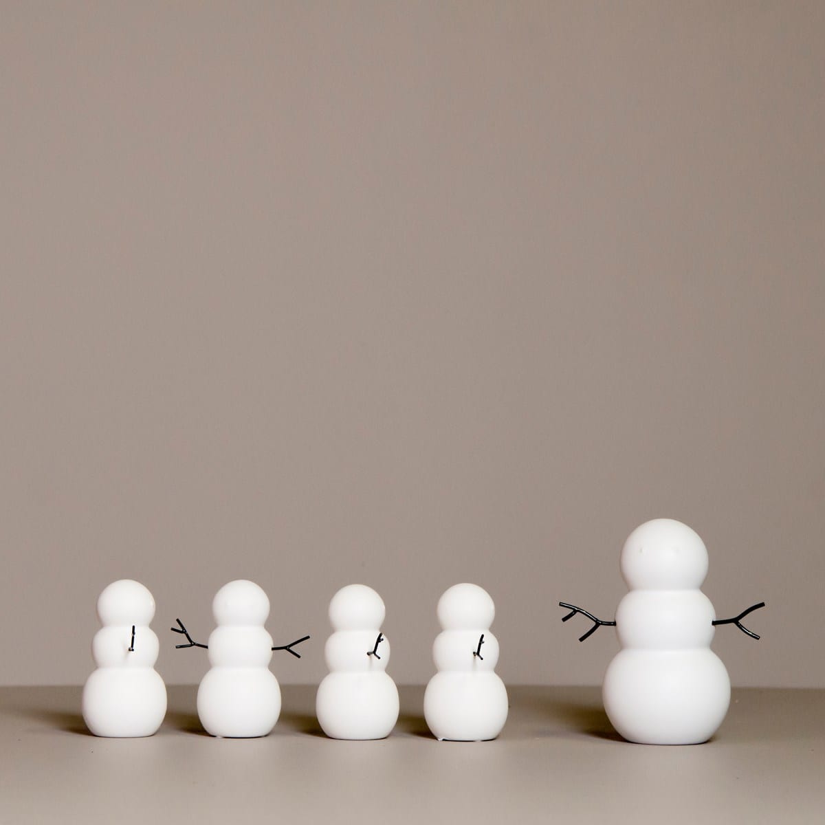 SNOWMAN features four minimalist snowman figures of different sizes in a row, each crafted from smooth white spheres with black stick arms, evoking curated décor vibes against a plain beige background. Gate21.ee