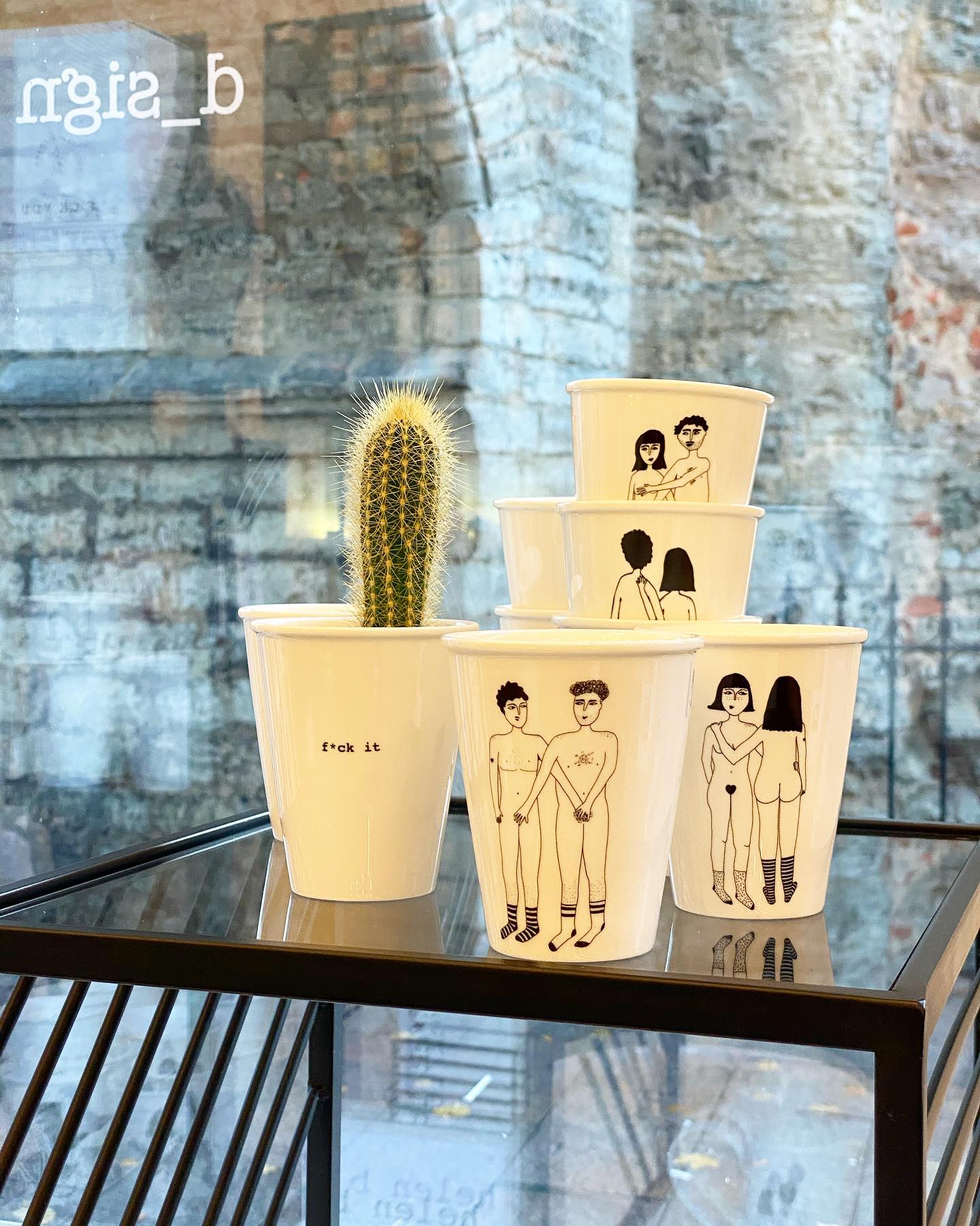 A white "4 Boobies Cup" featuring minimalist black line art sits stacked by a cactus in a "f*ck it" pot on a black shelf, with a stone wall visible through the window, evoking stylish Tallinn shopping vibes. Gate21.ee