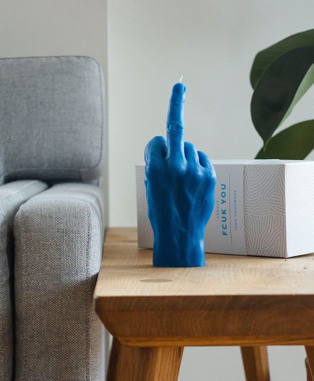 The F*ck You Candle - Blue, shaped like a hand making a middle finger gesture, rests on a wooden table beside a sofa and white patterned box. Ideal for Tallinn shopping or unique gifts from your favourite concept shop. Green plant in background. Gate21.ee