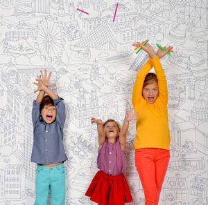Three children stand in front of a black-and-white illustrated cityscape wall at a curated concept shop. They face forwards with arms raised, wearing colourful clothes: blue, purple, and orange tops with matching trousers or skirt. Gate21.ee