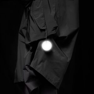 A person wearing a black, crinkled coat with the Dangle - Round - Grey accessory hanging from one pocket, posed against a black background—ideal inspiration for concept shop or gift shop displays in Tallinn shopping districts. Gate21.ee