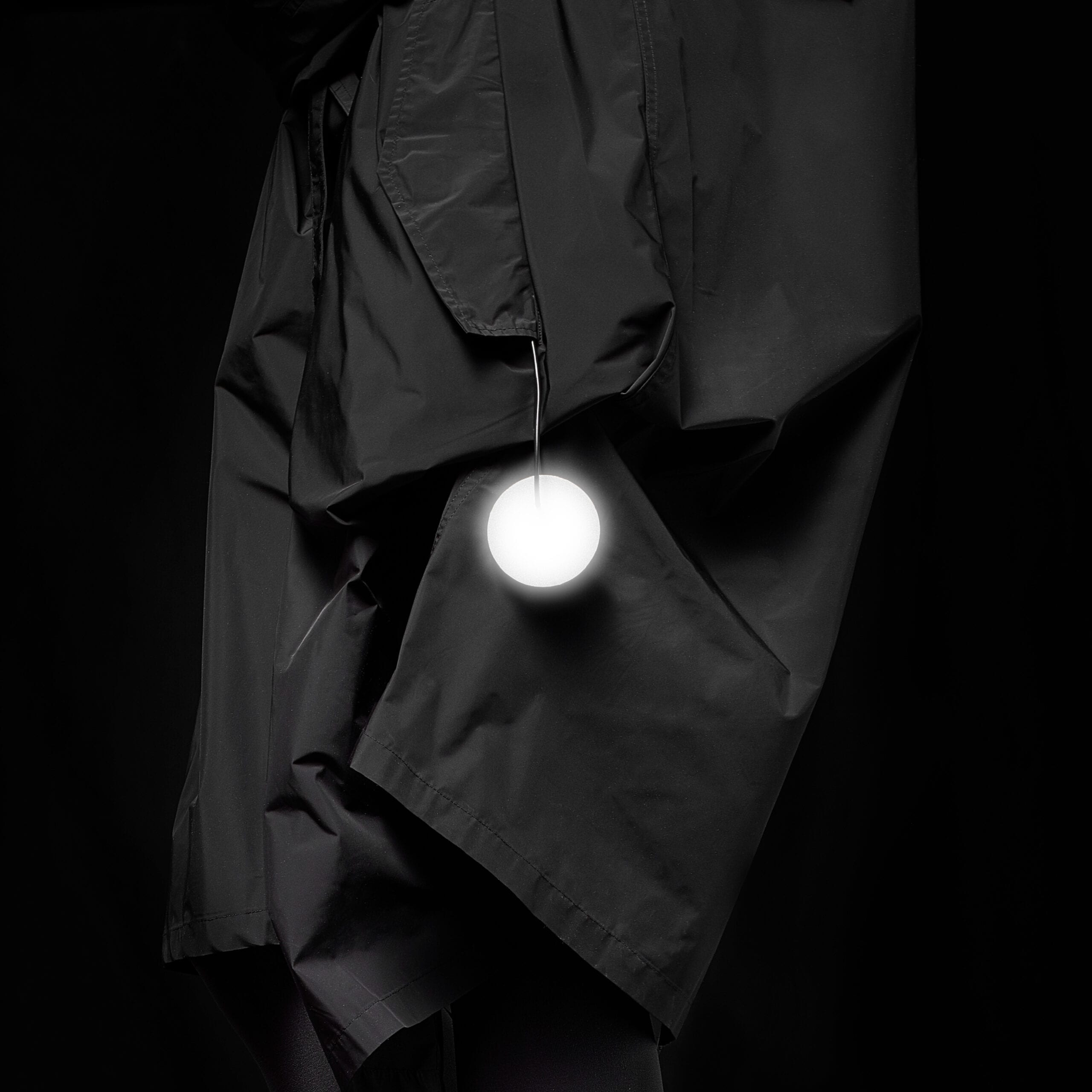 A person wearing a black, crinkled coat with the Dangle - Round - Grey accessory hanging from one pocket, posed against a black background—ideal inspiration for concept shop or gift shop displays in Tallinn shopping districts. Gate21.ee