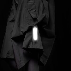 A person wearing a black coat with a Dangle - Round - Grey light attached stands against a solid black background, reflecting the sleek minimalism found in Estonian design and Tallinn’s concept shops. Gate21.ee