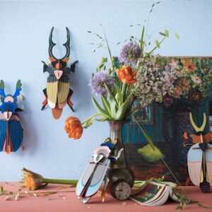 A vase with orange and purple flowers sits on a table near a painting and three colourful Giant Stag Beetle wall decorations—unique Estonian design, ideal for concept stores or gift shops. Gate21.ee