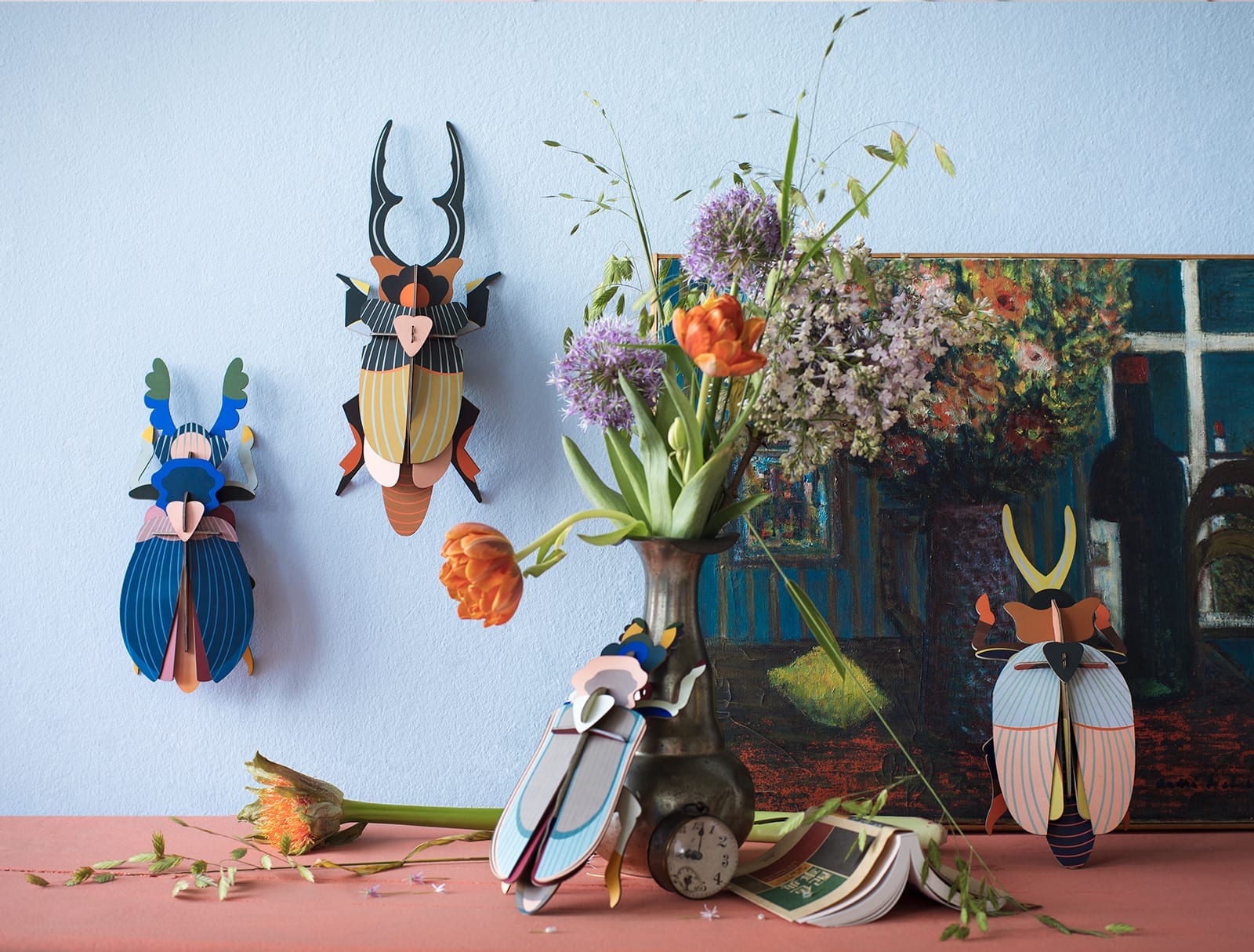 A vase with orange and purple flowers sits on a table near a painting and three colourful Giant Stag Beetle wall decorations—unique Estonian design, ideal for concept stores or gift shops. Gate21.ee