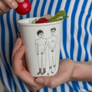 A person in a blue and white striped shirt holds the 2 Willies Cup, a white mug showcasing Estonian design with two nude figures in socks. The cup, filled with radishes, gets an extra one added by another hand. Gate21.ee