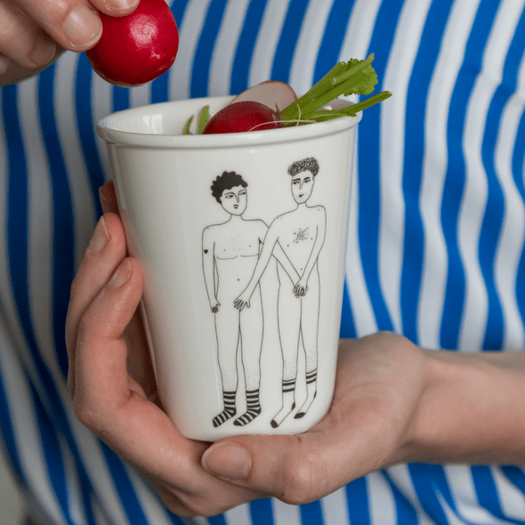 A person in a blue and white striped shirt holds the 2 Willies Cup, a white mug showcasing Estonian design with two nude figures in socks. The cup, filled with radishes, gets an extra one added by another hand. Gate21.ee