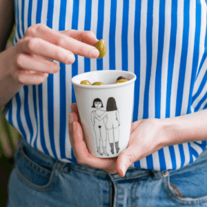 A person in a blue and white striped shirt and blue jeans holds the "4 Boobies Cup," a cup featuring an illustration of two women, one lifting an olive from a cup of green olives—a charming example of Estonian design. Gate21.ee