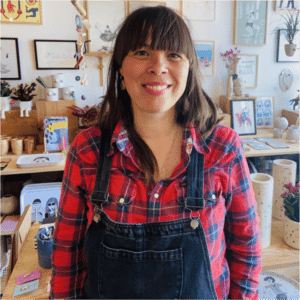 A woman with long brown hair and a fringe, wearing a red checked shirt and dark denim dungarees, smiles while standing in a concept shop displaying art, pottery, framed pictures, and the 4 Boobies Cup on tables and walls. Gate21.ee