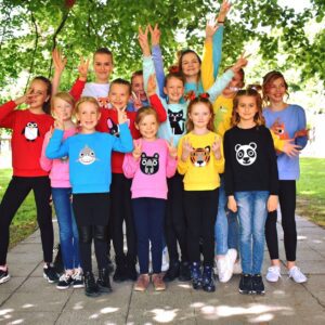 Thirteen children pose outside on a paved path, smiling and making peace signs. They’re wearing colourful Kids Dino jumpers with playful animal faces, surrounded by vibrant green trees and grass—ideal for those seeking unique concept shop ideas. Gate21.ee