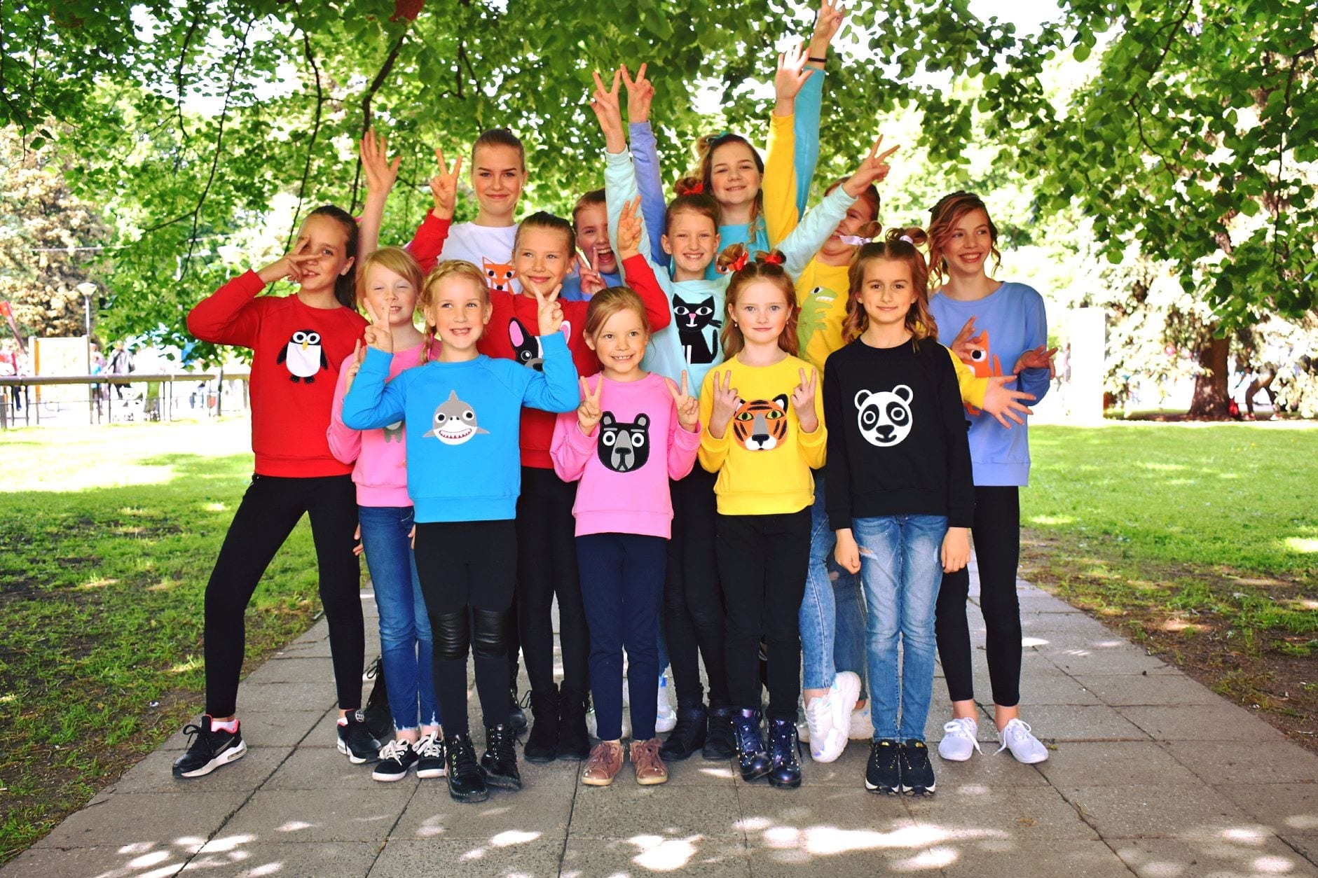 Thirteen children pose outside on a paved path, smiling and making peace signs. They’re wearing colourful Kids Dino jumpers with playful animal faces, surrounded by vibrant green trees and grass—ideal for those seeking unique concept shop ideas. Gate21.ee