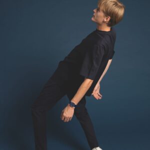 A person with short blond hair, dressed in black with white trainers and a Navy Blue / Silver / 40mm watch, leans back against a dark blue backdrop, capturing the essence of Estonian design and originality. Gate21.ee