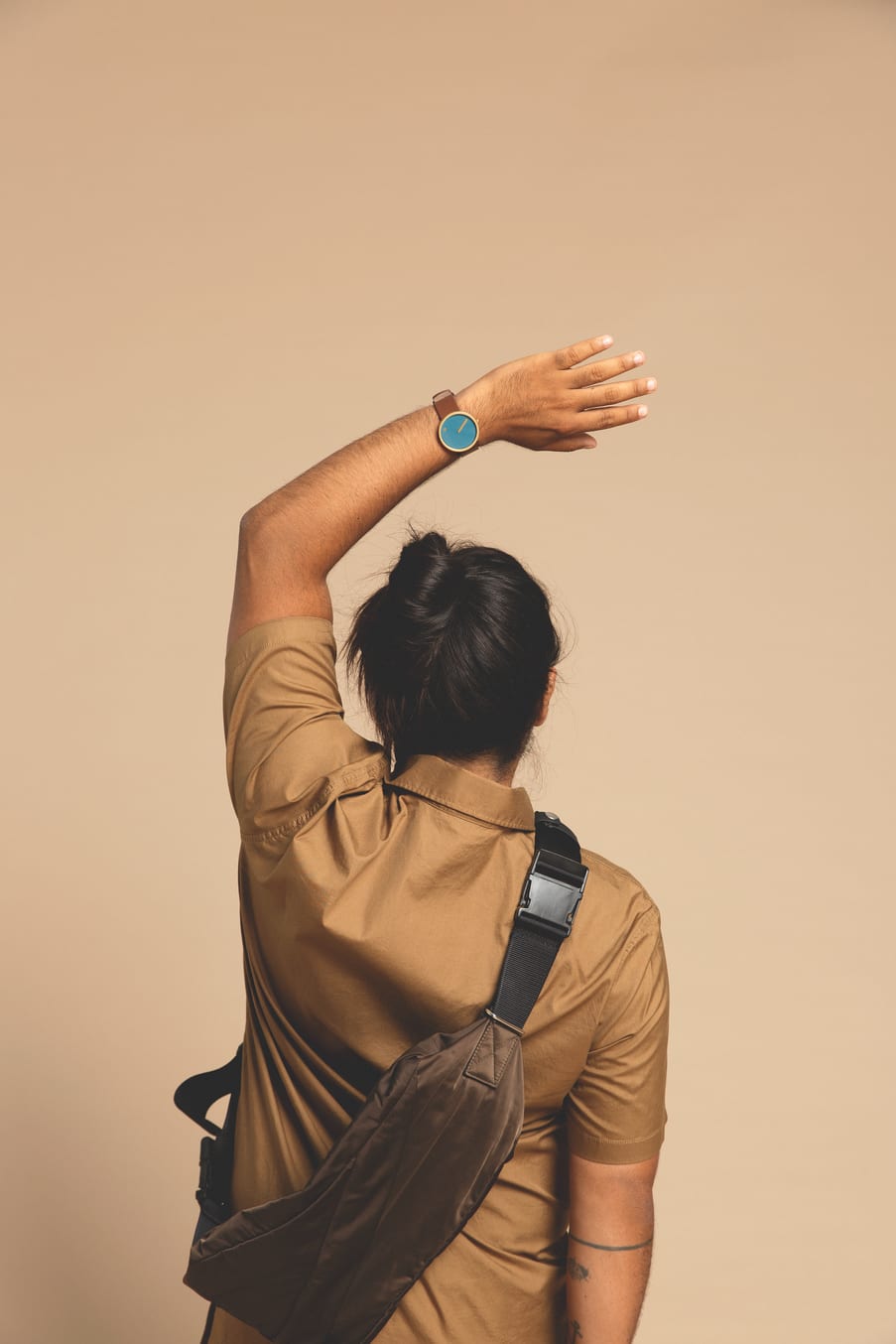 A person in a brown shirt and crossbody bag, hair tied up, stands with their back to the camera against a beige backdrop. They raise an arm, showing off the Dusty Blue / Gold / 40mm watch—ideal inspiration for Tallinn shopping or concept visits. Gate21.ee