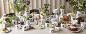 A table is set with mugs featuring black-and-white Estonian design illustrations, each holding various fruits, vegetables, plants, and flowers. Plates with snacks and cakes are also arranged among the mugs and floral displays. Gate21.ee