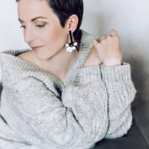 A person with short dark hair wearing a chunky light grey jumper, curated statement earrings featuring black and white circular elements&mdash;perfect for an Estonian design&ndash;inspired gift shop&mdash;sits with arms crossed, looking down and to the side. Gate21.ee