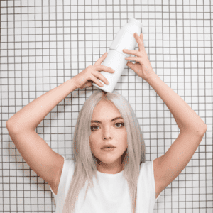 A person with long, straight, platinum blonde hair and a white shirt stands before a white tiled wall, balancing the Closca Wave - White bottle on their head—showcasing the innovative displays in Tallinn’s shopping and concept shops. Gate21.ee
