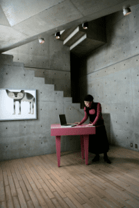 A person stands at a pink desk with a laptop in a modern concrete room, reminiscent of a Tallinn shopping concept store. The wall features minimalist cat artwork and geometric stairs with spotlights above, set on light wooden plank flooring. Gate21.ee