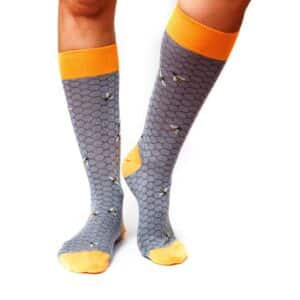 A person wears Bees - Regular knee-high grey socks featuring a honeycomb pattern, small bee designs, and yellow accents—a unique choice for curated style from your favourite concept store. Gate21.ee