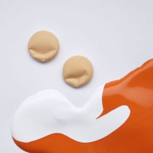 Two round, beige VITAMIN - K&Ouml;IK objects rest on a white surface beside bold splashes of white and orange paint&mdash;a curated, elegant presentation reminiscent of Estonian design found in select gift shops. Gate21.ee