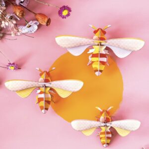 Three paper craft bees with yellow, orange, and white stripes are arranged on a pink background, circling a yellow centre. Dried flowers and petals in the upper left corner add charm—perfect for unique ideas in any gift shop display. Gate21.ee