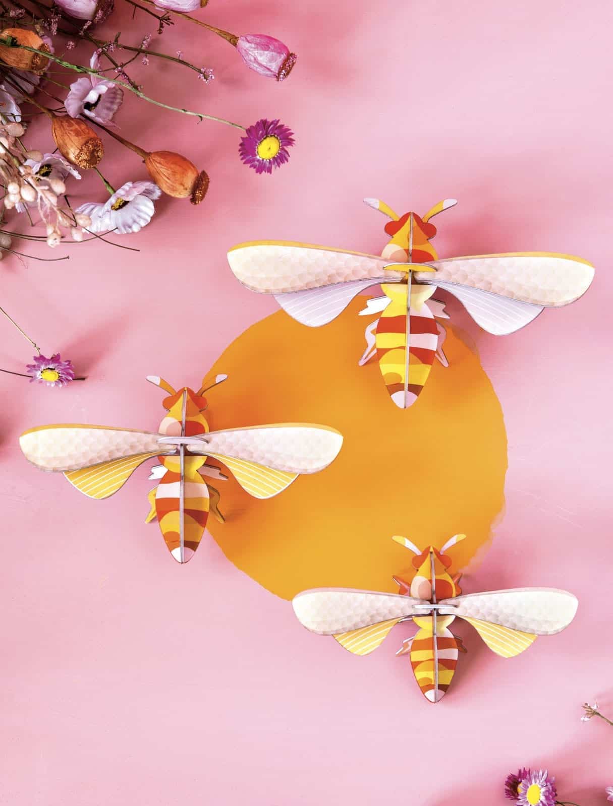 Three paper craft bees with yellow, orange, and white stripes are arranged on a pink background, circling a yellow centre. Dried flowers and petals in the upper left corner add charm—perfect for unique ideas in any gift shop display. Gate21.ee