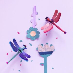 Curated papercraft dragonflies in blue and red rest on a light purple background, surrounded by simple paper flowers and scattered pink and blue confetti—perfect inspiration for any creative gift shop or Tallinn shopping adventure. Gate21.ee