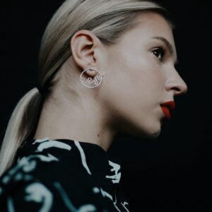 A woman with blonde hair in a low ponytail wears silver Abstract Vibes earrings and a black and white patterned top—her bold red lipstick completes her striking Estonian-inspired style. Gate21.ee