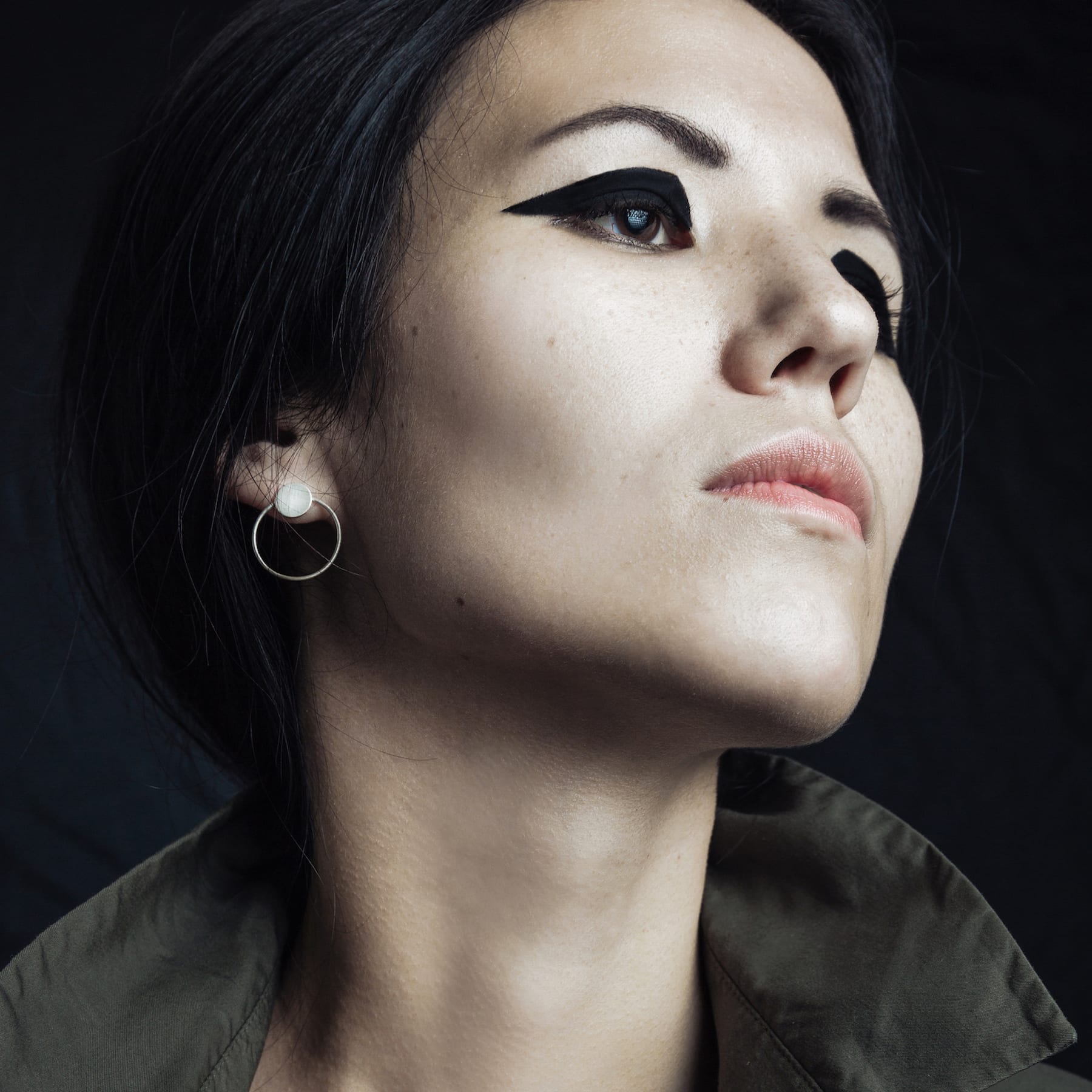 A woman with dark hair and bold black eyeliner looks upward in a concept store, wearing a green jacket and Circles on the Water - Silver earrings, inspiring unique Tallinn shopping ideas against a dark background. Gate21.ee