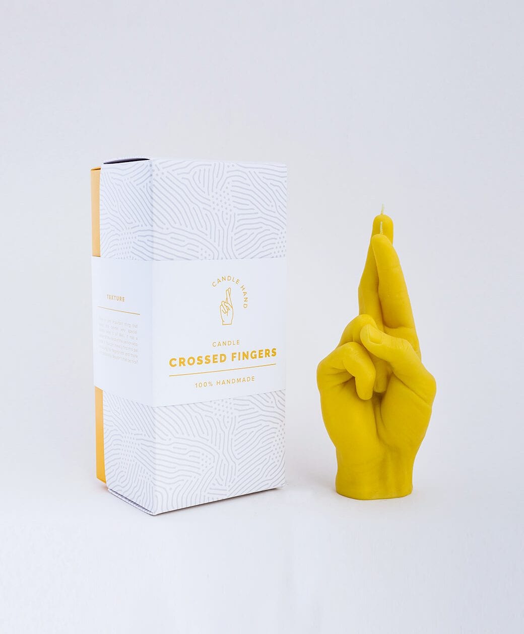 A yellow Fingers Crossed - Yellow candle, shaped like a hand with crossed fingers, sits beside its white box—an eye-catching 100% handmade gift or a unique find for your Tallinn shopping adventure. Gate21.ee