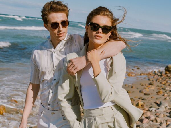 Two people in light-coloured outfits and sunglasses stand on a rocky beach, inspiring unique ideas for summer style. One has their arm around the other's shoulders as the ocean and sky stretch out behind them. Gate21.ee