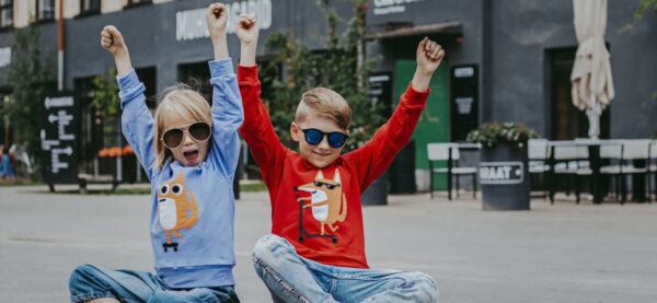 Two kids sit on the pavement with arms raised, wearing sunglasses, jeans, and Kids Doggy cartoon animal print sweatshirts. Outdoor tables and plants create a fun backdrop—perfect inspiration for Tallinn shopping family outings. Gate21.ee