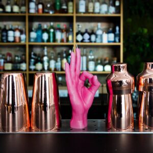 A row of copper cocktail shakers lines the bar counter with OK - Pink, a hand-shaped sculpture making an OK gesture, at the centre—showcasing the unique finds available when shopping in Tallinn. Shelves of liquor bottles fill the background. Gate21.ee