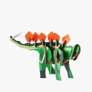 The Stegosaurus paper model, inspired by Estonian design, has a green and dark green body, orange back plates, and two pointed tail spikes. It is displayed against a plain white background. Gate21.ee