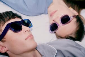Two people wearing curated sunglasses lie on a white surface, facing upwards. One sports blue-rimmed shades, the other light purple. Both are dressed in light-coloured clothing, embodying unique ideas in style. Gate21.ee