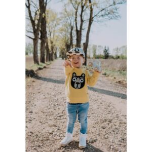 A young child wearing the Kids Bear yellow bear-face sweatshirt, blue jeans, white shoes, and a chequered cap stands on a tree-lined dirt path, holding a wooden toy car from an Estonian gift shop and pointing forward. Gate21.ee