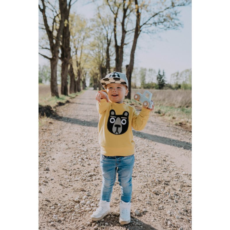 A young child wearing the Kids Bear yellow bear-face sweatshirt, blue jeans, white shoes, and a chequered cap stands on a tree-lined dirt path, holding a wooden toy car from an Estonian gift shop and pointing forward. Gate21.ee