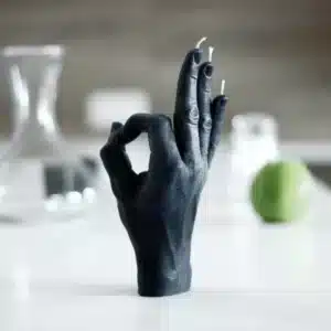 The OK - Black is a black sculpted hand in an “OK” gesture with candle wick fingertips, standing upright on a white surface. Ideal for concept shops or Tallinn shopping, it’s displayed with a glass container and blurred green object behind. Gate21.ee