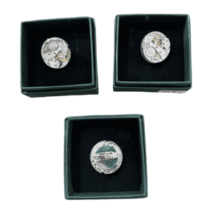 Three silver-coloured Clock pins, featuring Estonian design, are displayed in individual small square dark green boxes with black interiors, arranged in a triangular layout on a white background. Gate21.ee