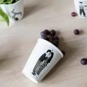 A white paper cup with a black illustration of two people standing close together, lying on its side on a light wooden surface next to scattered grapes—perfect for a curated concept shop or stylish gift shop. Other cups and a green plant are partially visible. Gate21.ee