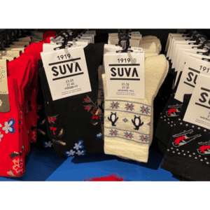 Several pairs of colourful patterned socks are displayed hanging on hooks, with SUVA brand tags and sizes visible. Perfect for a gift shop, these curated designs feature flowers, birds, and guitars in vibrant red, black, cream, and blue. Gate21.ee