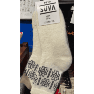 A pair of off-white wool socks with a black snowflake pattern near the top, hanging with a tag labelled 1919 SUVA and size information, displayed in a concept store celebrating Estonian design. Gate21.ee
