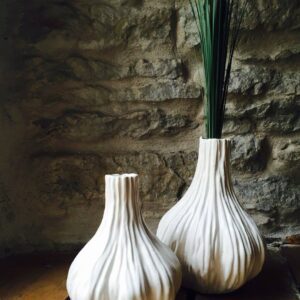 The Paper vase, featuring a white organic design inspired by garlic bulbs, sits on a wooden floor with tall green stems. This stylish piece is perfect for any home and makes an ideal find for Tallinn shopping or concept shops. Gate21.ee