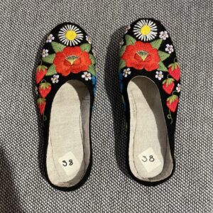 A pair of black slip-on shoes with brightly coloured floral embroidery, featuring curated Estonian design, are placed side by side on a grey textured surface. Size labels 38 are inside each shoe&mdash;perfect for a thoughtful gift shop find. Gate21.ee
