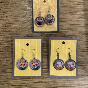 Three pairs of animal-themed earrings are displayed on yellow patterned cards against a wooden surface. These unique ideas feature illustrations of a bat, fox, and pig, each pair with a metal hook for wearing. Gate21.ee