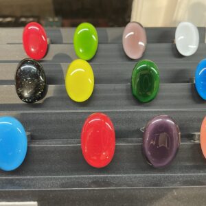 A curated display of twelve INGA KOPPEL glass rings, each oval-shaped with smooth, polished stones in vibrant solid colours, arranged in four rows on a black stand&mdash;an elegant showcase of Estonian design for your concept shop. Gate21.ee