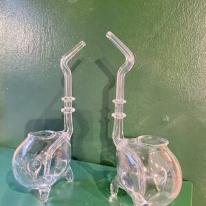 Two identical SYLI earrings with a round base, three legs, and long curved necks are displayed on a green surface against a green wall. Their clear glass form highlights curated Estonian design with an artistic and unusual structure. Gate21.ee